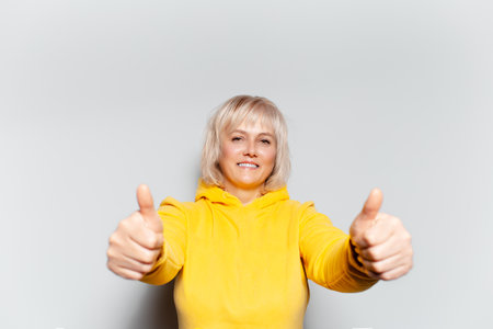 Studio portrait of happy blonde woman showing thumbs up on white background, wearing yellow sweater.