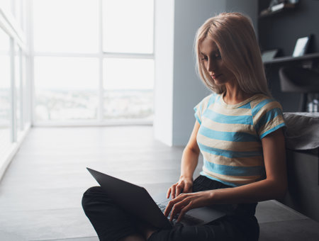 Portrait of blonde girl sitting on the floor while using the laptop on background of panoramic...