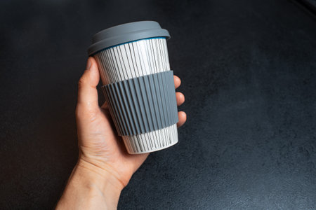Close-up of male hand holding a ceramic cup for coffee takeaway on black background.