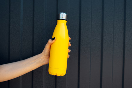 Close-up of female hand holding thermo bottle of yellow color on background of black metal fence.の写真素材