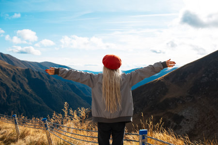 Back view of happy blonde girl on peak of the mountains, wearing grey jacket and red beret.