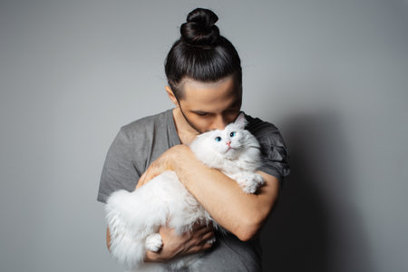 Studio portrait of young man with white cat in arms while kissing the pet on grey background.の写真素材
