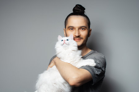 Studio portrait of young man with white cat in arms on grey background. Pet with blue eyes.の写真素材
