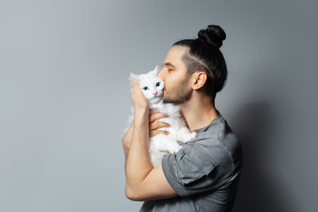 Studio portrait of young man with white cat in arms while kissing the pet on grey background.の写真素材