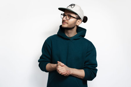 Young guy with hair bun looking away on white background. Wearing hoodie, cap and eyeglasses.