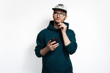 Thoughtful young guy with fingers on chin, holding smartphone in hand. Isolated on white background.