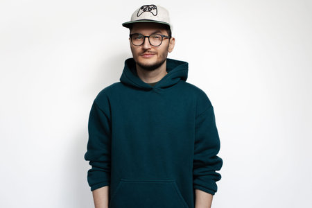 Young man isolated on white background, wearing eyeglasses, cap and green hoodie sweater.