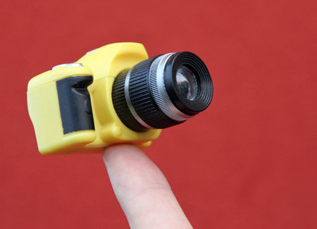 index finger holding a small yellow suspended camera and red background