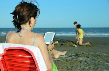 tanned woman reads the ebook on the seashore in the summer while her children playing on the beach