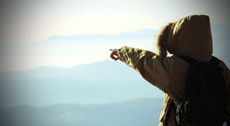 young Explorer with winter jacket with hood and black backpack indicates the horizon from above the top of the mountainsの写真素材
