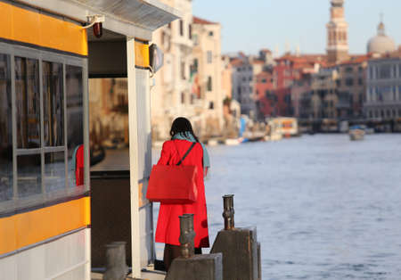 elegant young woman dressed in red with red purse awaits boat on the Grand Canal in Venice Italyの写真素材