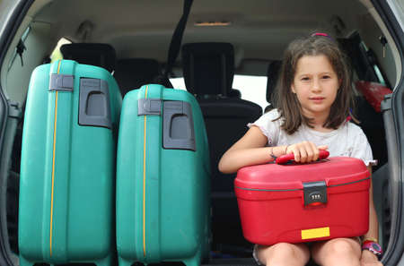 beautiful little girl with brown hair long loads the suitcase in the trunk of the car already full of suitcases before leaving for the holidaysの写真素材