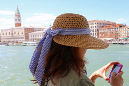 young woman with long brown hair and a wide straw hat uses the smartphone to send a picture you have just taken in Venice in Italyの写真素材
