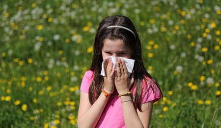 Young girl with long brown hair with allergy to the pollen blows her nose with the handkerchief in the middle of the meadowの写真素材