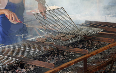 cook with a blue apron cooking meat burgers on the grill of a fireplace at a festival