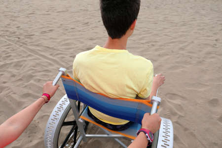 young boy on the special wheelchair on the beach in summer and the arms of his sisterの写真素材