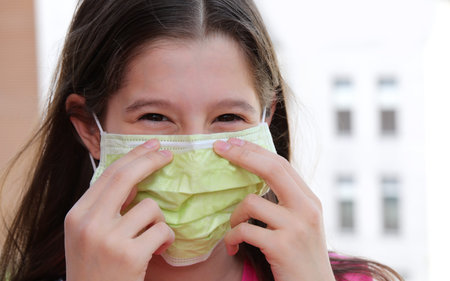 little girl with smiling eyes and without worries fixes the green surgical mask for protection from...