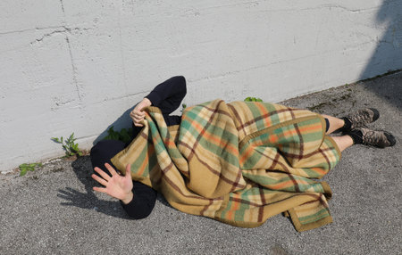 young homeless man lying on the street on the asphalt under an old blanket and his hand raised to...