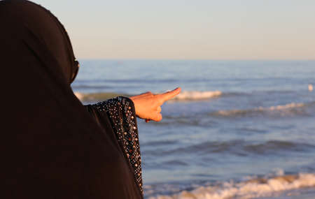 Arab woman with veil on her head pointing to a distant point in the sea at sunsetの写真素材