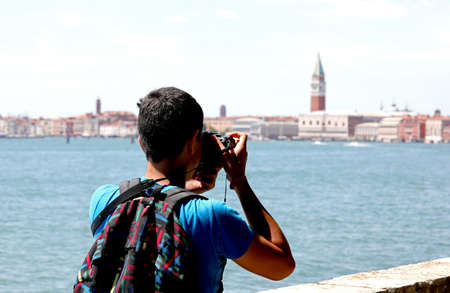 young photographer with backpack on his shoulders while taking a photograph at the bell tower of San Marco in Venice in Italyの写真素材