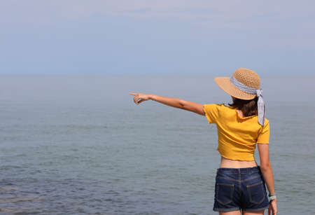 young girl in yellow shirt and jeans by the sea in the summer points to a point towards the horizonの写真素材