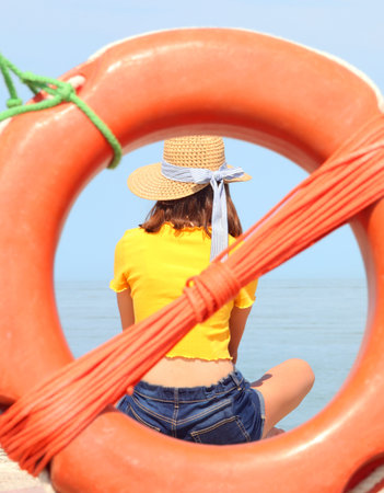 young girl with yellow shirt and wide straw hat inside an orange lifebuoy by the seaの写真素材