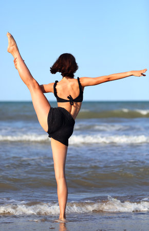 young woman by the sea performs difficult balance exercises with raised legの写真素材