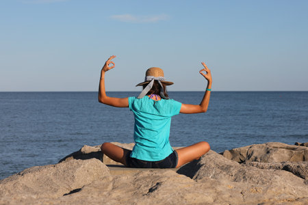 young slender girl with blue t-shirt does meditation and yoga exercises by the sea on the rocksの写真素材