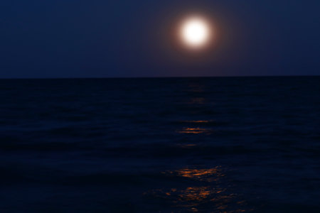 full moon rising from the sea with reddish effect that is reflected on the waterの写真素材