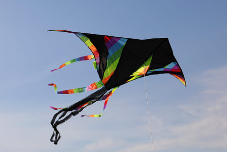 Large black kite with multicolored rainbow stripes flies high in the blue sky tied to a string