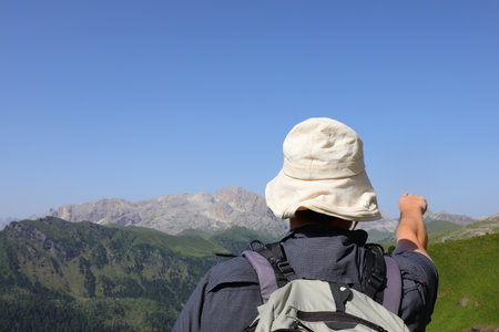 Elderly hiker with a white sun hat on his head pointing towards a mountain peakの写真素材