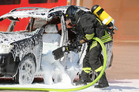 Firefighter with oxygen tank and protective helmet spraying white extinguishing foam to put out the fire that broke out in the destroyed car after the road accidentの写真素材