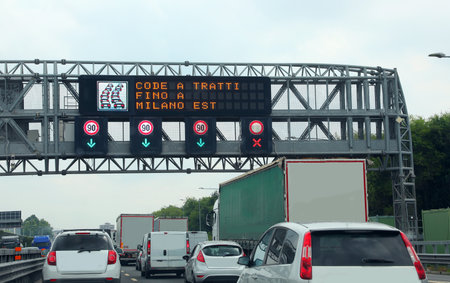 Portal with a message on the motorway with the text that means Queues A Tratti up to MILAN EAST for heavy traffic with many vehicles on the roadsの写真素材