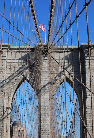 Iconic view of NYC with Waving American Flag on the top of Tower of Brooklyn Bridge and many steel...