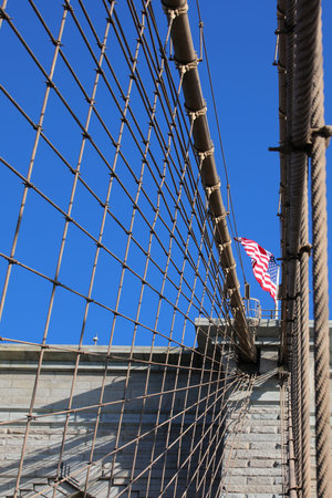 huge united states flag waving amidst the steel suspension wires of the brooklyn bridge in new york with blue heavens behindの写真素材