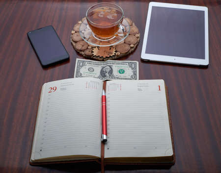 On the wooden table is a plate, a cup of tea, a cell phone, Address Book, Pen, Dolar, moneyの写真素材