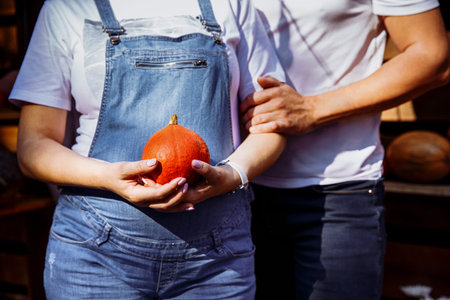 husband and pregnant wife in denim suits hug each other, she holds a pumpkin in her hands.