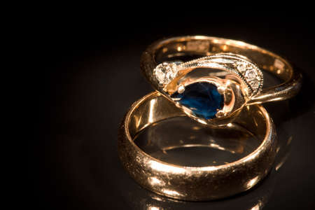 Two golden rings with sapphire and diamonds isolated on black with reflection, light painting, full frame camera shotの写真素材