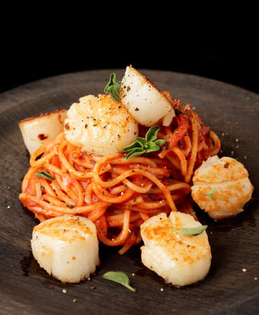 Pasta with fried scallops, oregano and tomato sauce on wooden plate isolated on black backgroundの写真素材