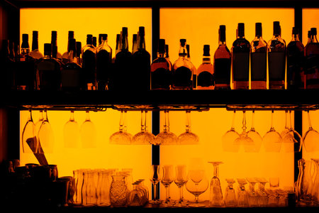 Bottles and glasses sitting on shelf in a bar, back lit, brand names removed