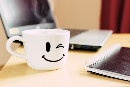 Smiling cup of coffee, notepad and laptop on the deskの写真素材