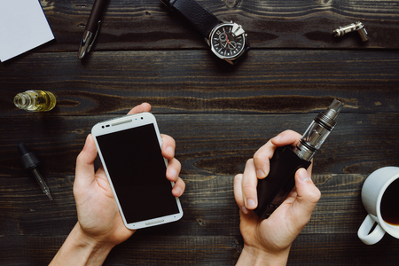 Man smoking vape or electronic cigarette and using smartphone. View from above. Vaping set, watch, coffee on the wooden background. Hipster or bussinesman style.の写真素材