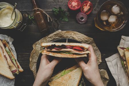 Hands holding fresh delicious club sandwich, sauces and drink on the wooden dark table, top view.の写真素材