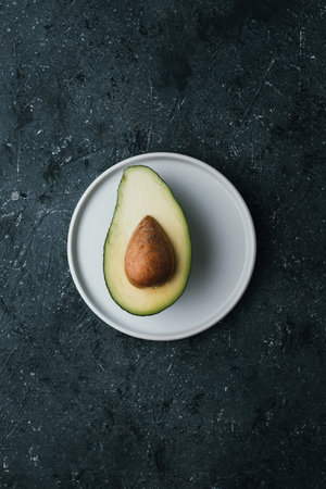 Ripe half avocado on white plate on dark background, top view