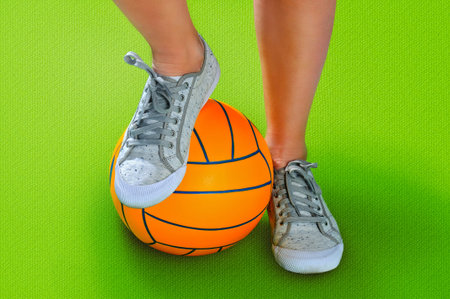 Female feet in white sneakers with an orange ball on a green background.