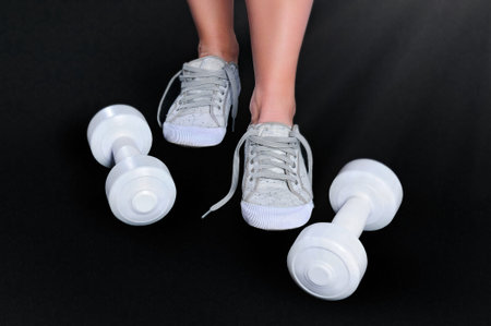 Female feet in white gym shoes and white dumbbells on a dark surface