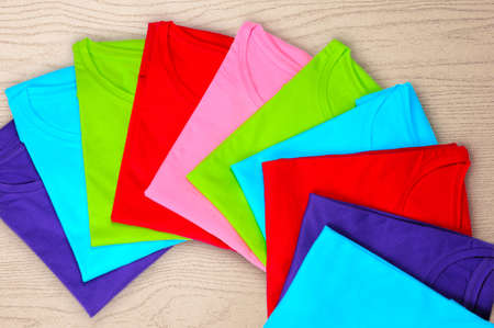 Turquoise, light blue, green, red, pink women's t-shirts lie on a wooden backgroundの写真素材