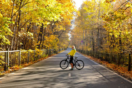 Slim caucasian girl in a yellow jacket poses with a bicycle on the road among the autumn forestの写真素材