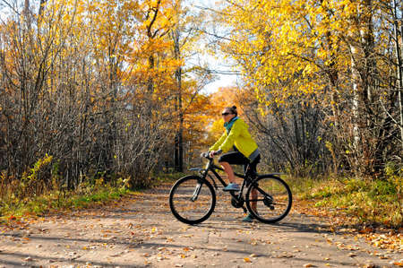 Slim caucasian girl in a yellow jacket poses with a bicycle on the road among the autumn forestの写真素材