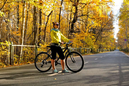 Slim caucasian girl in a yellow jacket poses with a bicycle on the road among the autumn forestの写真素材